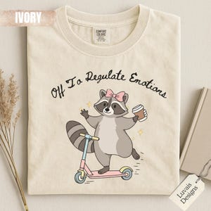 Puede incluir: Una camiseta color marfil con un gráfico de un mapache con un lazo rosa, montado en un patinete rosa y sosteniendo una taza de café. El texto sobre el mapache dice, Off To Regulate Emotions. La camiseta tiene cuello redondo y etiqueta.