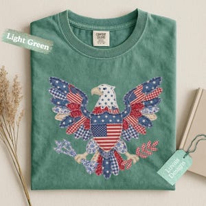 May include: A light green t-shirt featuring a central graphic of a bald eagle composed of various red, white, and blue patterned fabric patches. The eagle holds arrows and an olive branch, with a shield displaying the American flag on its chest.