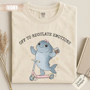 Op de afbeelding: Een ivoorkleurig t-shirt met een afbeelding van een blauwe vis met een kleine roze strik op een roze step, terwijl de vis een koffiebeker vasthoudt. De tekst boven de vis luidt "OFF TO REGULATE EMOTIONS" in zwarte letters. Comfort Colors label.