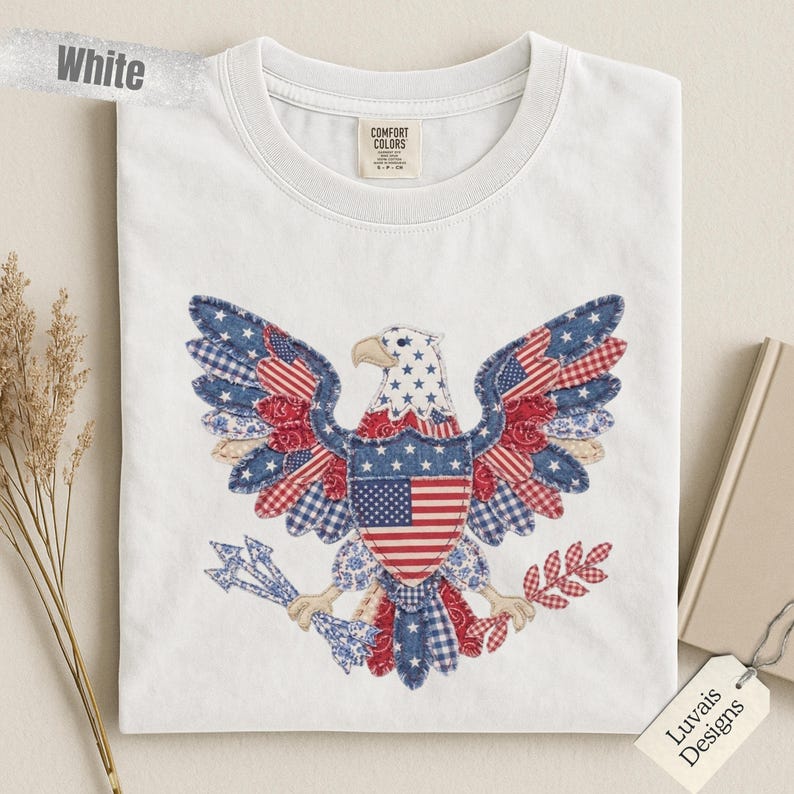 May include: A white t-shirt featuring a large, central graphic of a bald eagle designed with a patchwork of red, white, and blue patterns, including stars, stripes, and gingham. The eagle holds arrows in one talon and an olive branch in the other.