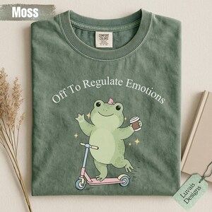 Puede incluir: Una camiseta de color verde musgo con el gráfico de una rana verde claro con un pequeño lazo rosa en la cabeza, montada en un patinete rosa mientras sostiene una taza de café. El texto sobre la rana dice Off To Regulate Emotions.
