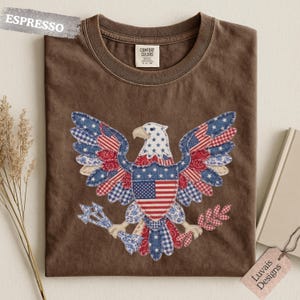 May include: An espresso brown t-shirt featuring a central graphic of a bald eagle composed of various red, white, and blue patterned fabric patches, including stars and stripes. The eagle holds arrows and an olive branch in its talons.