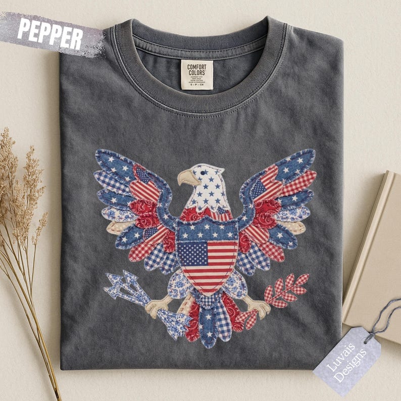 May include: A charcoal gray t-shirt featuring a large graphic of a bald eagle with wings spread. The eagle is designed with a patchwork pattern of red, white, and blue fabrics, including stars and stripes. The shirt has a Comfort Colors label.
