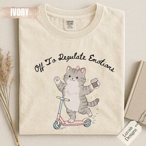 May include: An ivory t-shirt featuring a graphic of a grey tabby cat with a pink bow, riding a pink and blue scooter while holding a coffee cup. The text above the cat reads, Off To Regulate Emotions. The shirt has a crew neckline and short sleeves.