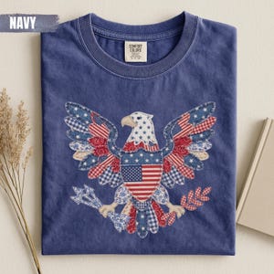 May include: A navy blue crew neck t-shirt featuring a large graphic of a bald eagle composed of various red, white, and blue patterned fabric patches. The eagle holds arrows and an olive branch, with a central shield displaying the American flag.