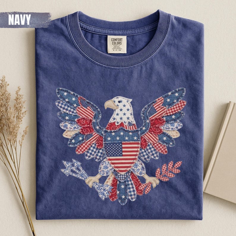 May include: A navy blue crew neck t-shirt featuring a large graphic of a bald eagle composed of various red, white, and blue patterned fabric patches. The eagle holds arrows and an olive branch, with a central shield displaying the American flag.
