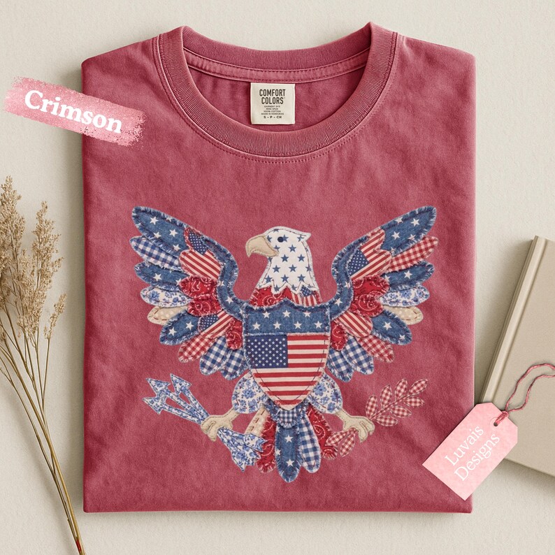 May include: A crimson crew neck t-shirt featuring a central graphic of a bald eagle composed of various red, white, and blue patterned fabric patches, including stars, stripes, and gingham, holding arrows and an olive branch in its talons.