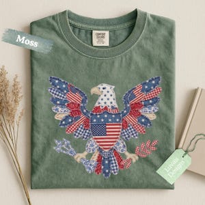 May include: A moss green crew neck t-shirt featuring a central graphic of a bald eagle composed of various red, white, and blue patterned fabric patches. The eagle holds arrows and an olive branch, with a prominent American flag design on its chest.