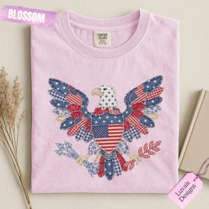 May include: A light pink crew neck t-shirt featuring a large graphic of a bald eagle with wings spread. The eagle is composed of various blue, red, and white patterned fabric patches, including stars and stripes. The word BLOSSOM is visible.