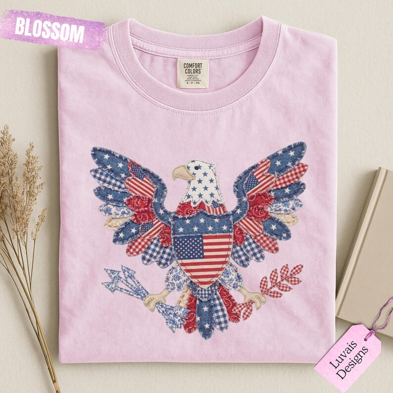 May include: A light pink crew neck t-shirt featuring a large graphic of a bald eagle with wings spread. The eagle is composed of various blue, red, and white patterned fabric patches, including stars and stripes. The word BLOSSOM is visible.
