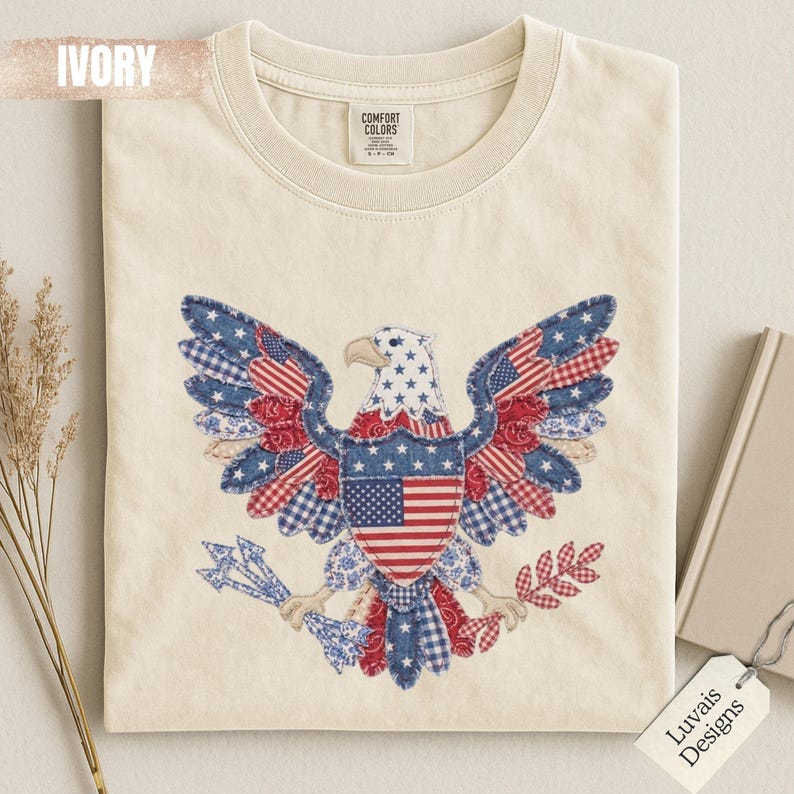 May include: An ivory t-shirt featuring a graphic of a bald eagle composed of various red, white, and blue patterned fabric patches. The eagle holds arrows and an olive branch, with a central shield displaying the American flag. The label reads Comfort Colors.