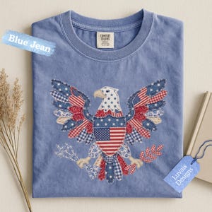 May include: A blue jean colored t-shirt featuring a large eagle graphic composed of various red, white, and blue patterned fabric patches. The eagle holds arrows and a branch, with a central shield displaying the American flag. The label reads Comfort Colors.