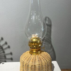 Puede incluir: Una lámpara de aceite de mimbre con una chimenea de vidrio transparente y una parte superior de metal dorado. La lámpara está sobre una superficie blanca.