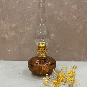 Może przedstawiać: Brązowa szklana lampa naftowa z przezroczystym szklanym kominem i złotym metalowym wierzchołkiem. Lampa stoi na jasnoszarym podłożu z gałązką żółtych kwiatów.