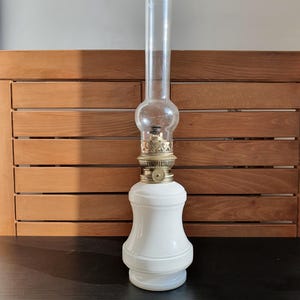 Puede incluir: Una lámpara de aceite de cerámica blanca con una chimenea de vidrio transparente y una base de latón. La lámpara tiene un diseño decorativo en la base y un cuerpo redondo de cerámica blanca.