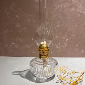 Może przedstawiać: Klara szklana lampa naftowa z pozłacanym metalowym wierzchołkiem i żebrowaną podstawą. Lampa stoi na białym tle z gałązką żółtych kwiatów.
