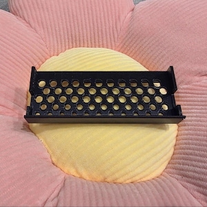 May include: A black, rectangular tray with numerous circular perforations, resting on a yellow and pink flower-shaped cushion. The tray has a raised edge and appears to be made of plastic or metal. The cushion has a ribbed texture.