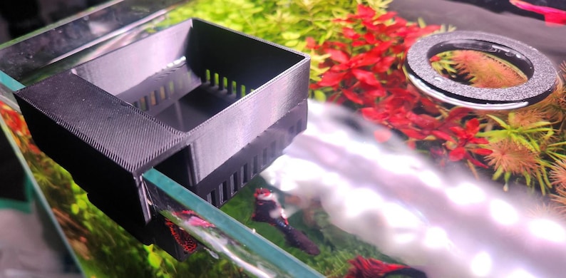 May include: A black plastic filter for an aquarium. The filter has a rectangular shape with a grid pattern on the front and a small opening on the side. The filter is attached to the side of the aquarium.