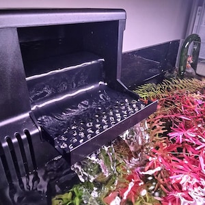 May include: A black aquarium filter with water cascading into a tank. The tank contains vibrant red and green aquatic plants. The filter has a stepped design, and the water flows over a textured surface.