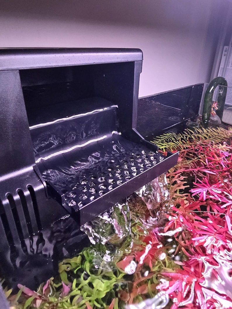 May include: A black aquarium filter with water cascading into a tank. The tank contains vibrant red and green aquatic plants. The filter has a stepped design, and the water flows over a textured surface.