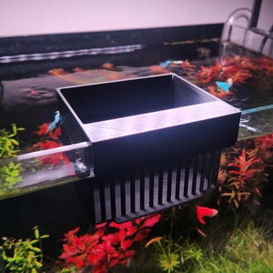 May include: A black plastic aquarium filter with a grid-like design hangs on the edge of a fish tank. The tank is filled with water and contains plants and fish.