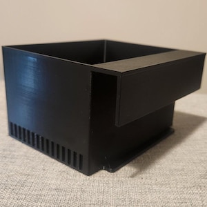 May include: A black, rectangular, 3D-printed object with a unique design. It features a square box shape with a protruding shelf on one side and a series of vertical slots on the front. The object is made of a matte black material.