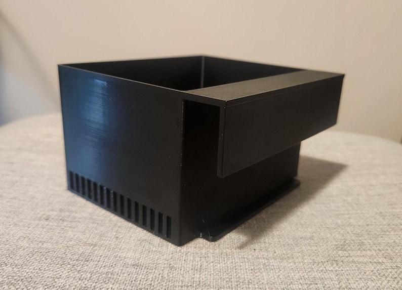 May include: A black, rectangular, 3D-printed object with a unique design. It features a square box shape with a protruding shelf on one side and a series of vertical slots on the front. The object is made of a matte black material.