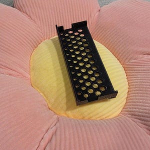 May include: A black, rectangular, perforated metal object with multiple circular holes. The object is resting on a flower-shaped cushion with pink petals and a yellow center.