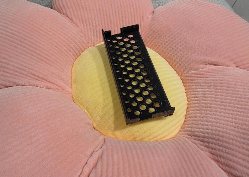 May include: A black, rectangular, perforated metal object with multiple circular holes. The object is resting on a flower-shaped cushion with pink petals and a yellow center.