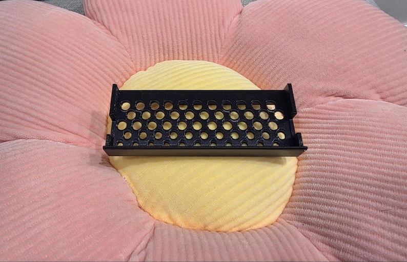 May include: A black, rectangular tray with numerous circular perforations, resting on a yellow and pink flower-shaped cushion. The tray has a raised edge and appears to be made of plastic or metal. The cushion has a ribbed texture.