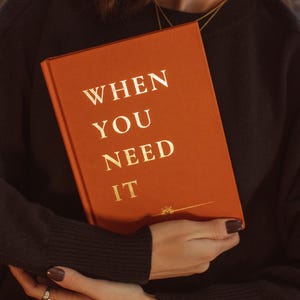 May include: A person is holding a book with an orange cover and gold lettering that reads "WHEN YOU NEED IT".