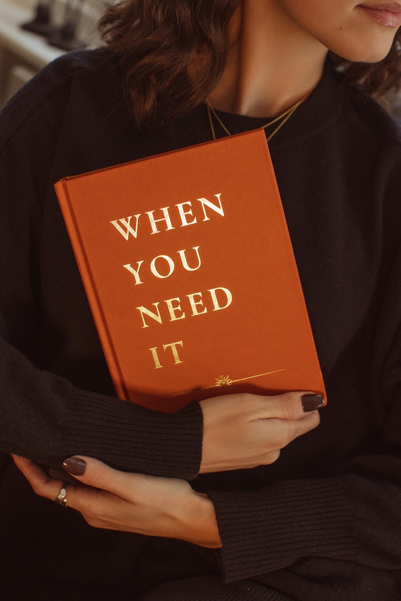 May include: A person is holding a book with an orange cover and gold lettering that reads "WHEN YOU NEED IT".