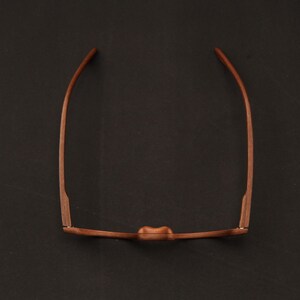 May include: A pair of brown wooden eyeglasses viewed from above, showing the bridge, the front frame, and the two temples extending outward. The frame has a smooth, natural wood grain texture and a curved bridge design for the nose.
