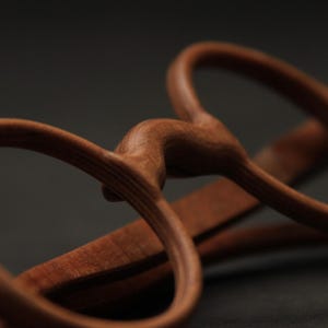 May include: A pair of eyeglasses crafted from brown wood, featuring rounded frames and a curved bridge. The natural grain of the wood is visible across the surface of the frames and the temples, which are positioned behind the front lens area.