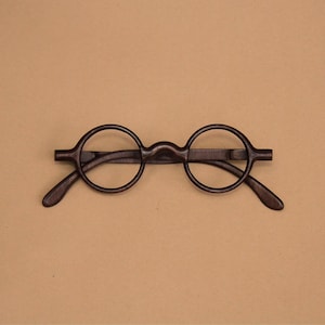 May include: A pair of dark brown wooden spectacles featuring round frames and a bridge that connects the two circular lenses. The temples extend outwards from the sides of the frames, showcasing a visible wood grain texture throughout the structure.