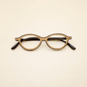 Puede incluir: Gafas de forma ovalada con una montura de veta de madera de color marrón claro con líneas horizontales visibles. Las varillas y los terminales están hechos de un material marrón oscuro sólido, creando contraste con los aros frontales de madera.