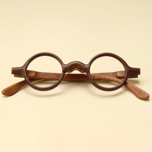 Peut inclure: Une paire de lunettes à monture ronde fabriquée en bois brun foncé. Les montures présentent un motif de grain distinct et un pont incurvé, avec des branches en bois brun plus clair s'étendant vers l'extérieur. Design symétrique.