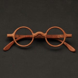 May include: Round wooden eyeglasses with a light brown finish. The glasses have a classic, minimalist design with circular lenses and slender arms. The frame is made of wood and rests on a dark background.