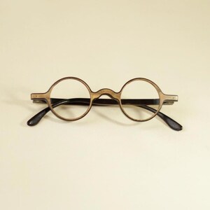 Puede incluir: Gafas redondas con una montura de madera de color marrón claro con un patrón de veta visible. Las gafas tienen un puente fino que conecta los aros circulares y patillas oscuras y delgadas que se extienden desde los lados de la montura.