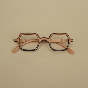 Puede incluir: Gafas con montura de madera de color claro y un borde marrón oscuro distintivo alrededor de los cristales cuadrados. Las patillas están hechas de la misma madera clara y el puente une ambos aros con un diseño suave y curvado.