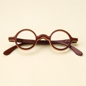 Puede incluir: Gafas redondas con montura y patillas de madera marrón oscuro. La montura tiene un acento de madera más claro alrededor de las lentes. Las gafas están sobre un fondo color crema.