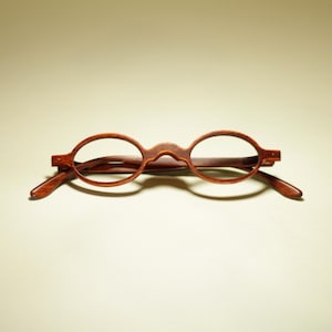 May include: A pair of round, brown eyeglasses with a bridge and temples. The glasses have a classic, vintage design, with a dark brown frame and lenses. The glasses are set against a neutral background.