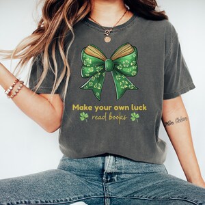 May include: A dark gray t-shirt with a green and gold book bow and the text "Make your own luck read books" with four shamrocks.
