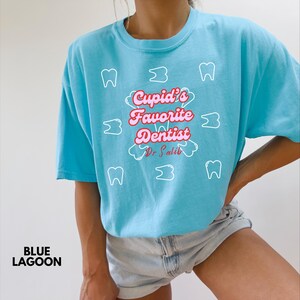 May include: A light blue t-shirt with the text "Cupid's Favorite Dentist Dr. Salib" in pink and white. The shirt also features white tooth and cloud graphics. The words "Blue Lagoon" are printed in the bottom left corner.