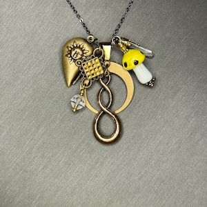 Puede incluir: Un collar dorado con múltiples colgantes. Los colgantes incluyen un corazón con un sol, un cuadrado con gemas amarillas, una seta, una cuenta de cristal y un símbolo de infinito. La cadena es de color gris oscuro.