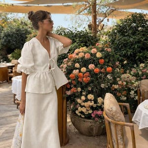 May include: A woman wearing a white linen two-piece outfit, consisting of a top with a V-neck and tie closure, and a matching skirt. The outfit is worn outdoors near a flower bed with orange, pink, and white flowers.