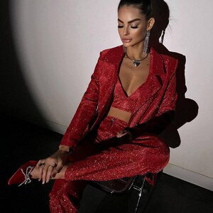 May include: A woman wearing a red sparkly blazer and matching pants. She is sitting on a chair and has her hair pulled back in a bun.