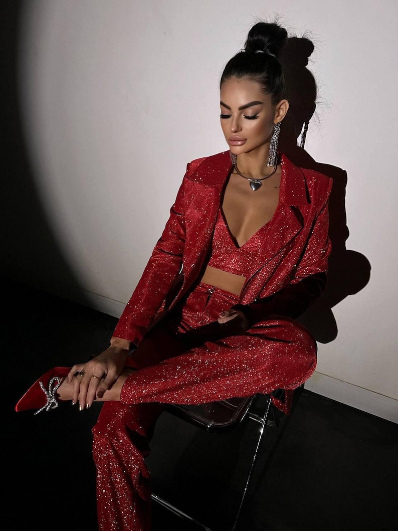 May include: A woman wearing a red sparkly blazer and matching pants. She is sitting on a chair and has her hair pulled back in a bun.