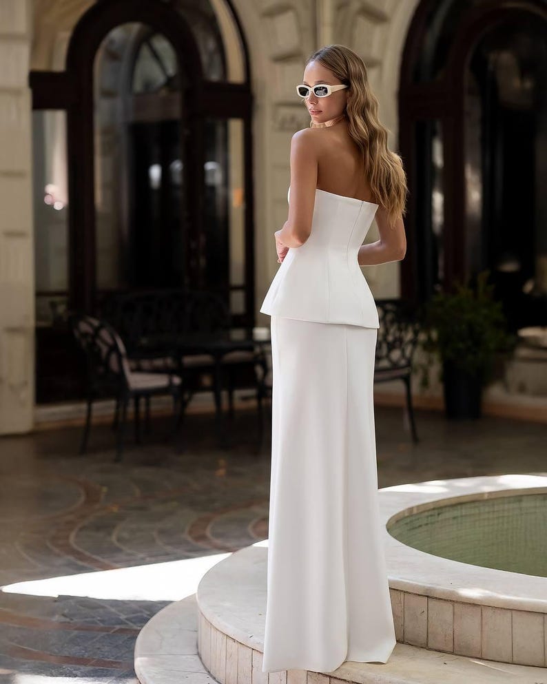 May include: A floor-length white strapless gown featuring a structured peplum waist and a straight skirt silhouette. The garment is paired with white-framed sunglasses, showcasing a clean, solid-colored design with a fitted bodice and flared peplum.