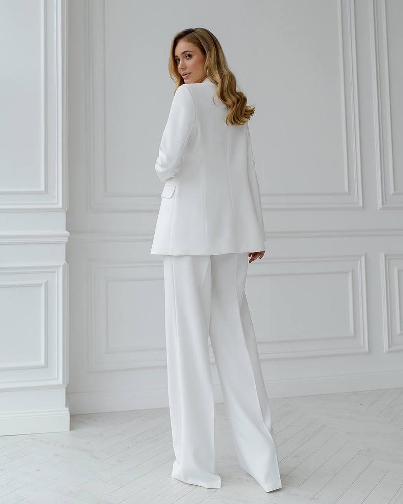May include: A white suit consisting of a blazer and wide-leg pants. The blazer has a classic design with lapels and pockets. The pants are long and flowing, creating a sleek, elegant silhouette. The suit is made of a smooth, solid white fabric.